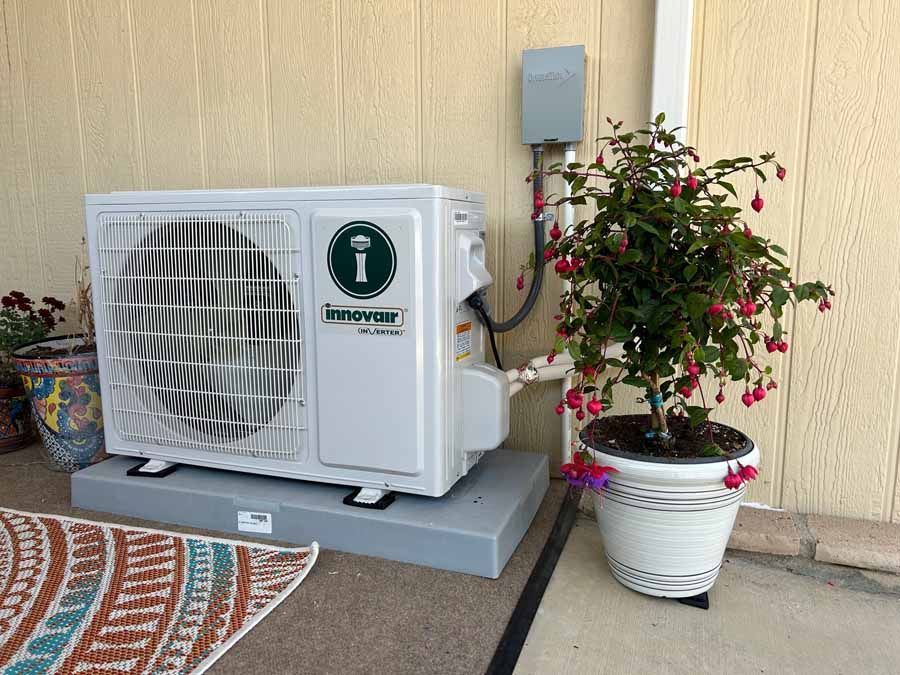 Outdoor air-conditioning unit beside a potted flowering shrub against a beige wall