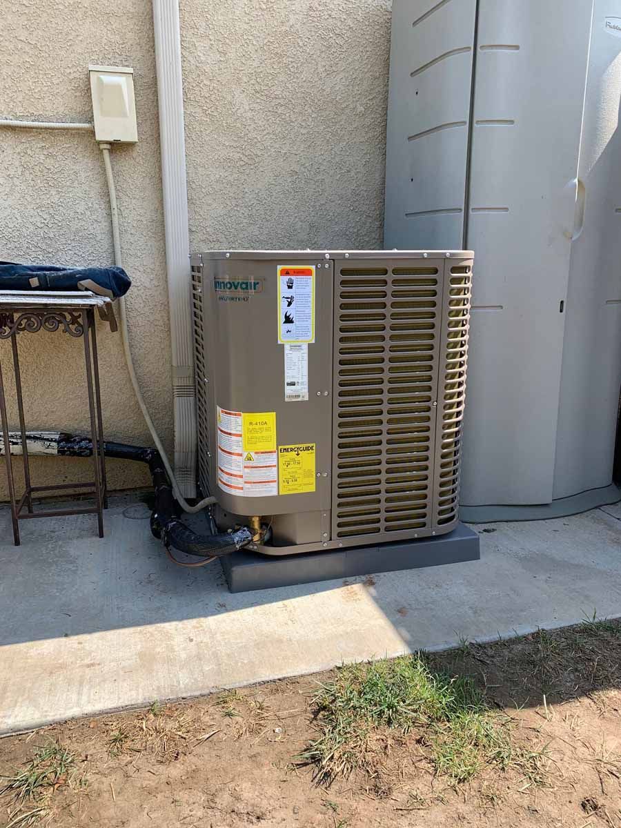 Outdoor air conditioning unit beside a stucco wall, with a chair and utility meter nearby.