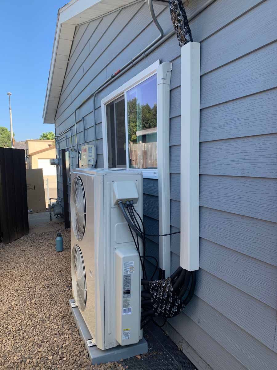 Outdoor wall with a mounted HVAC unit, electrical boxes, and white conduit on gray house siding.