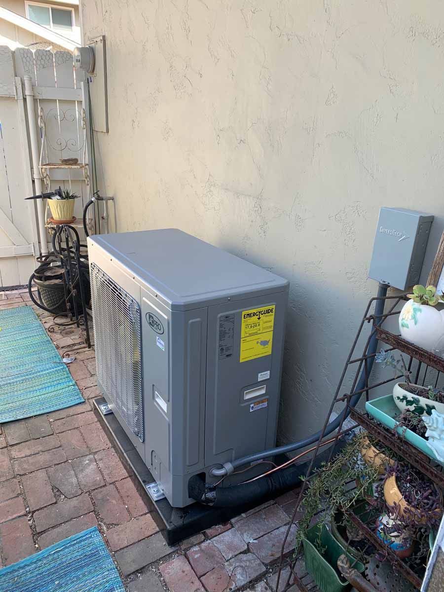 Outdoor HVAC condenser unit beside a brick walkway and house wall, with utility boxes and garden pots nearby
