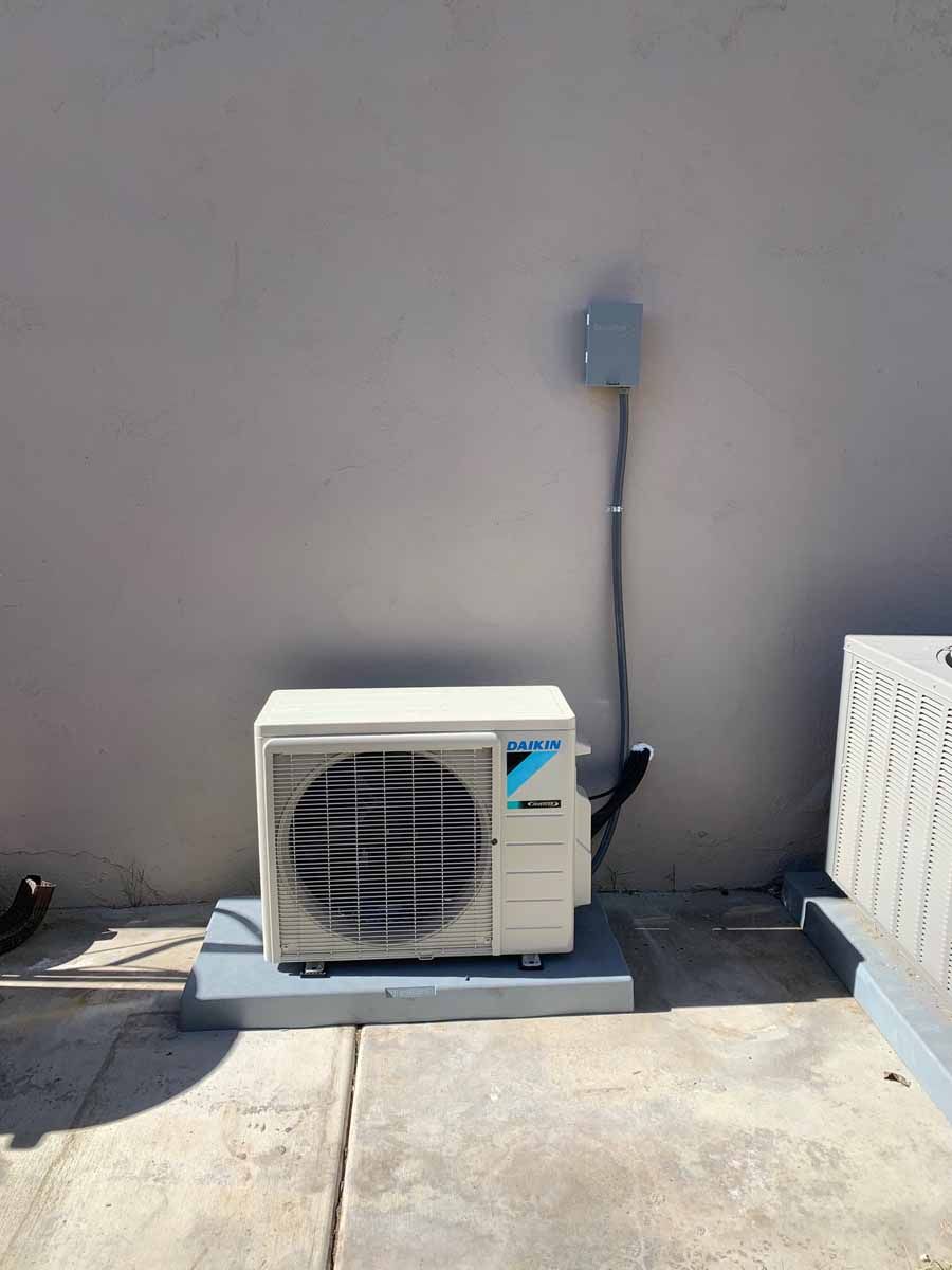 Outdoor air conditioning unit mounted against a gray wall on a concrete patio, with an electrical box above it