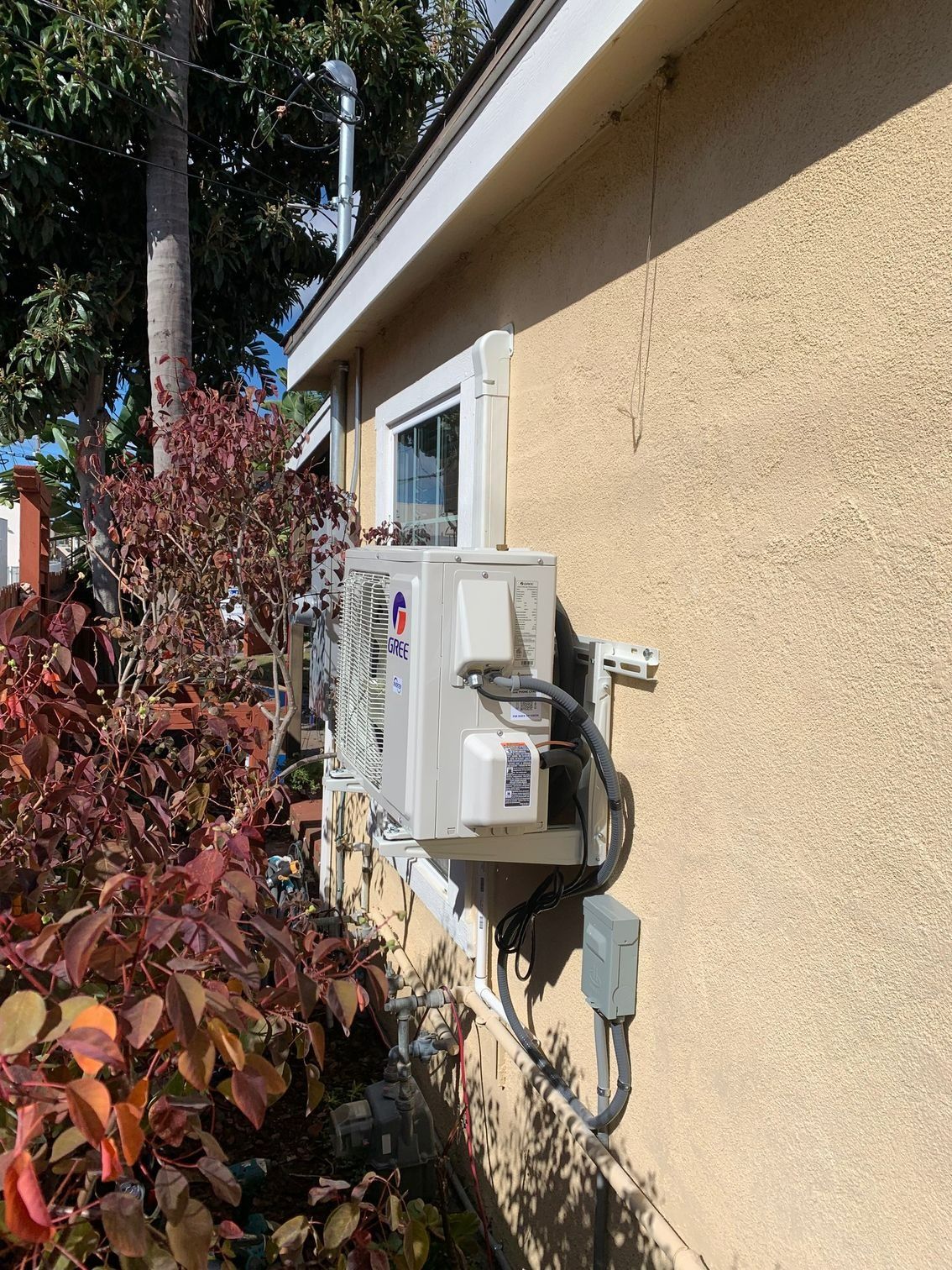 Outdoor wall-mounted air-conditioning unit beside a beige house, with red shrubs in the foreground.