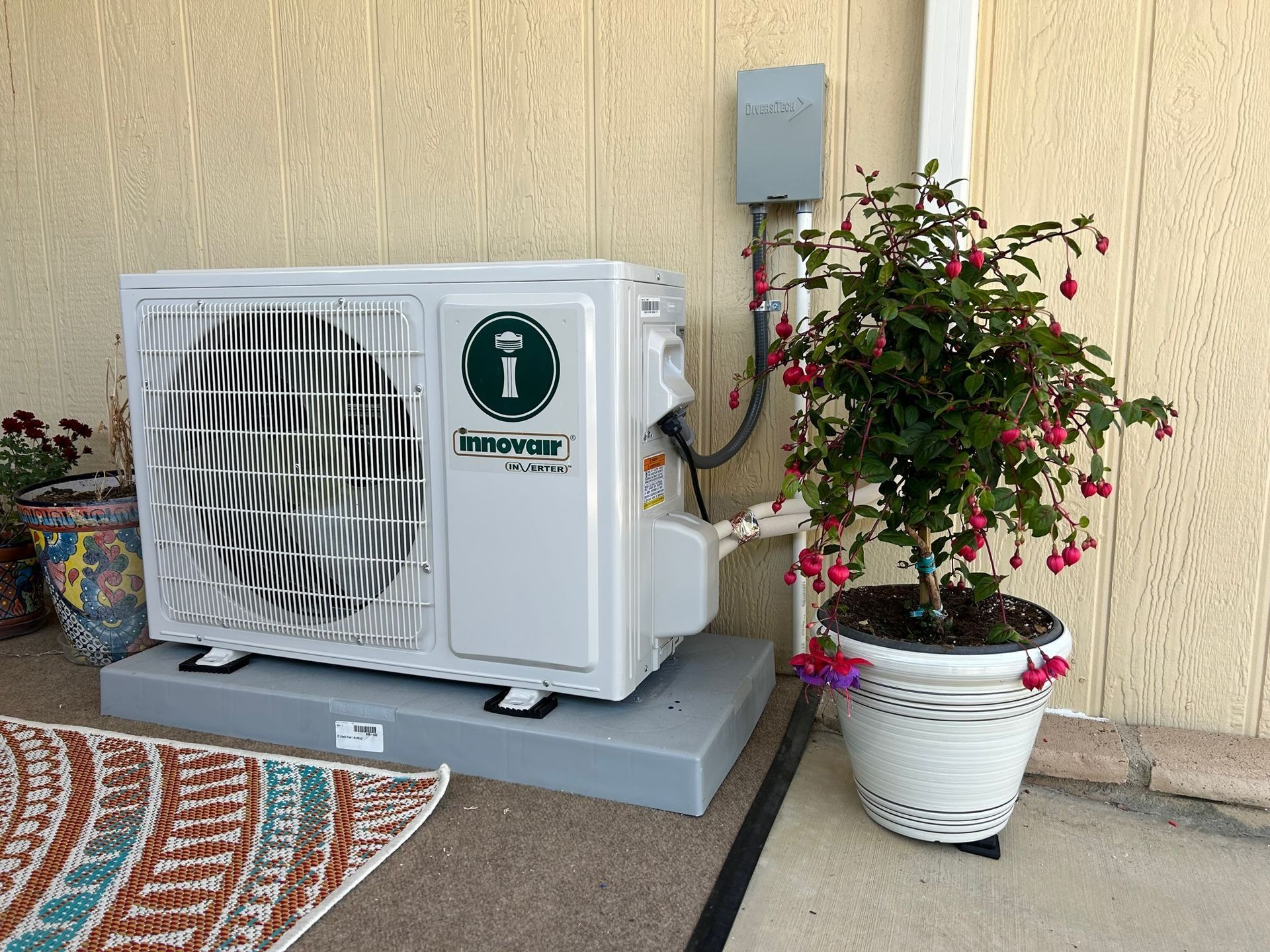 Outdoor air conditioner unit beside a potted flowering plant against a beige wall