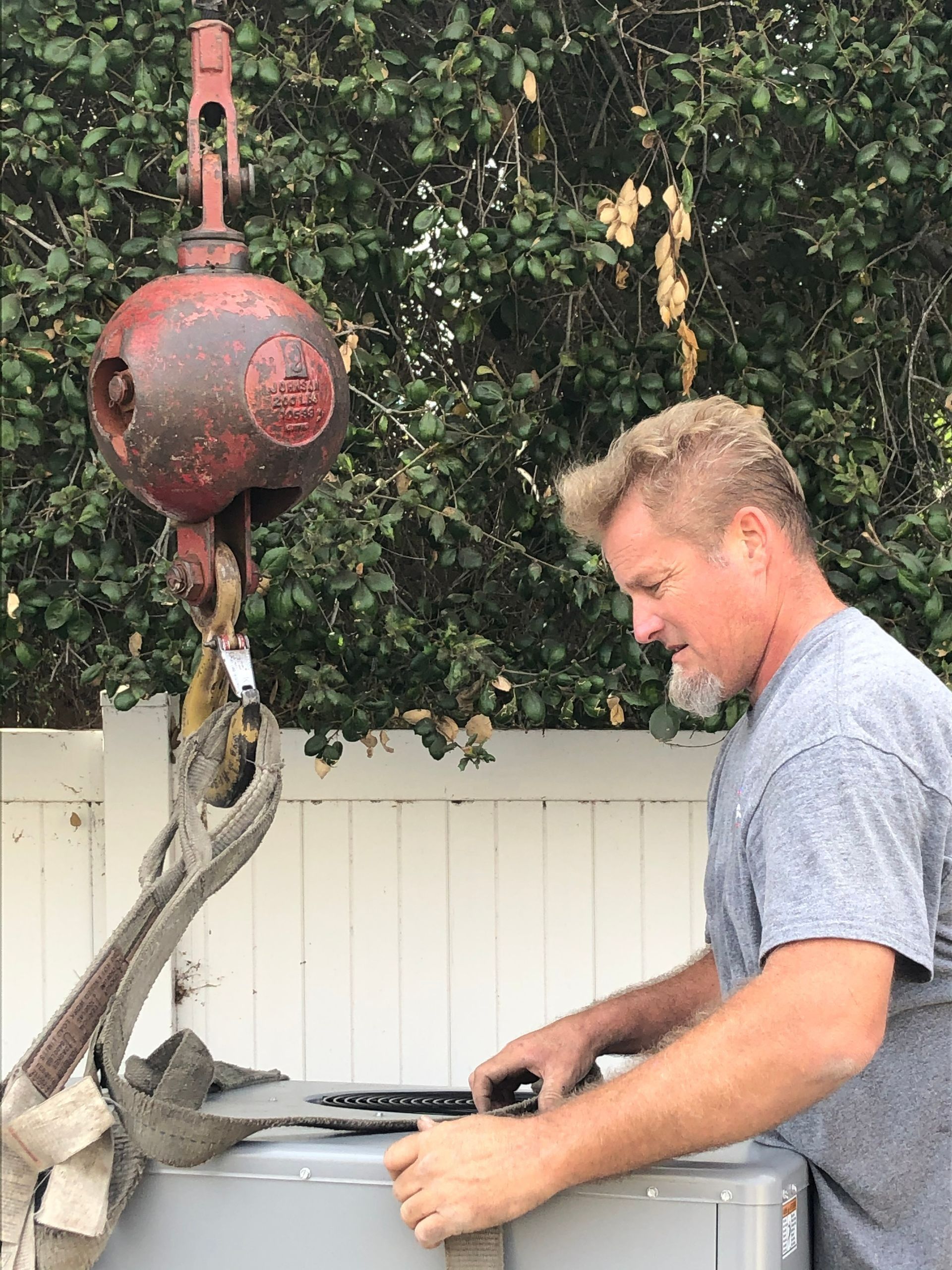 Man adjusting a hoist hook above a wrapped object outdoors beside a white fence and hedge