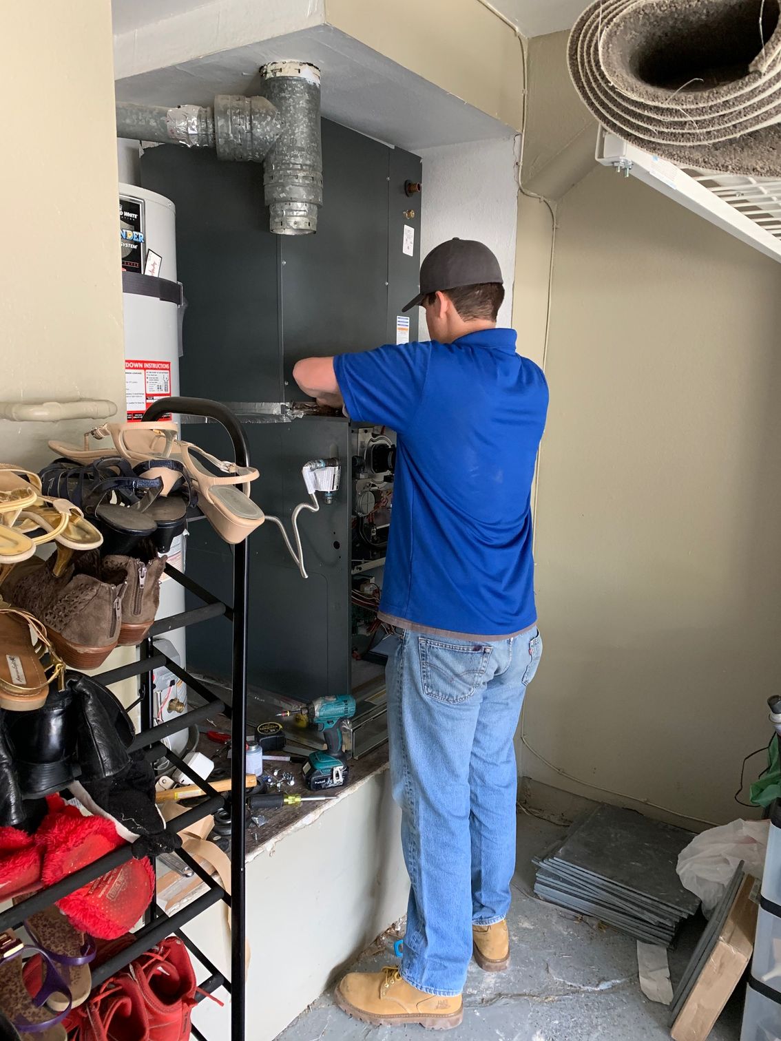 Technician in blue shirt working on a furnace in a utility closet, with tools and storage shelves nearby