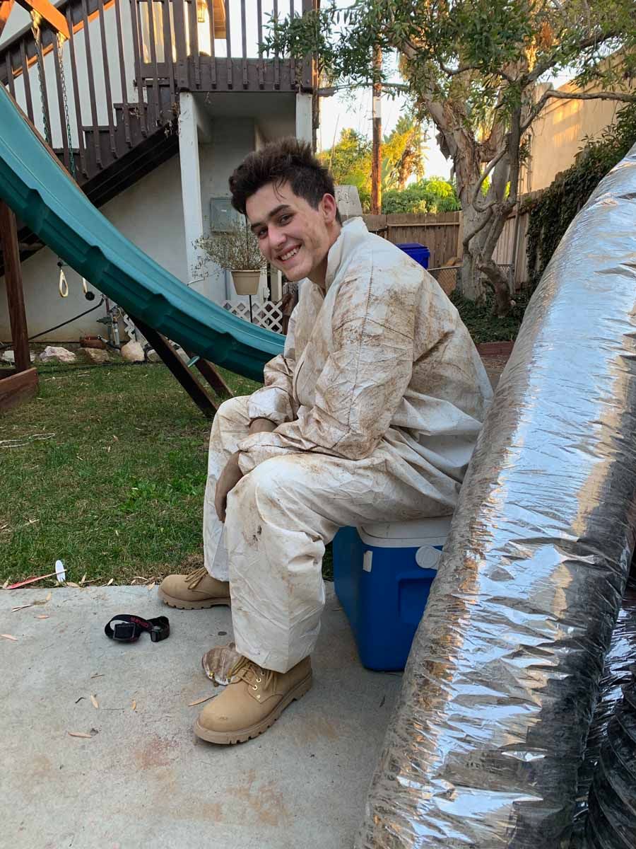 Person in a light outfit sitting on a blue cooler outdoors, smiling beside a playground slide.