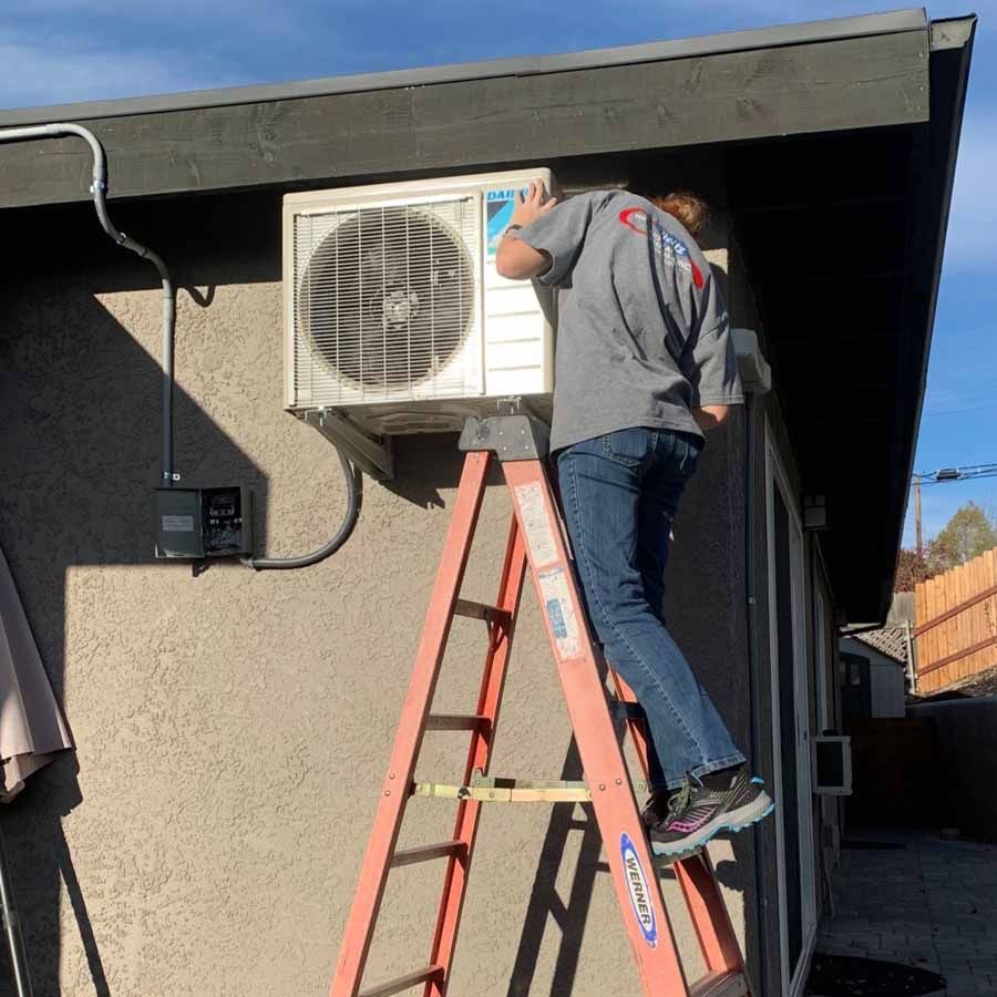 Technician on a ladder servicing an outdoor air conditioner unit on a house exterior.
