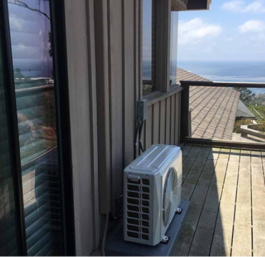 Air conditioner unit on a wooden deck beside a house, overlooking the ocean.