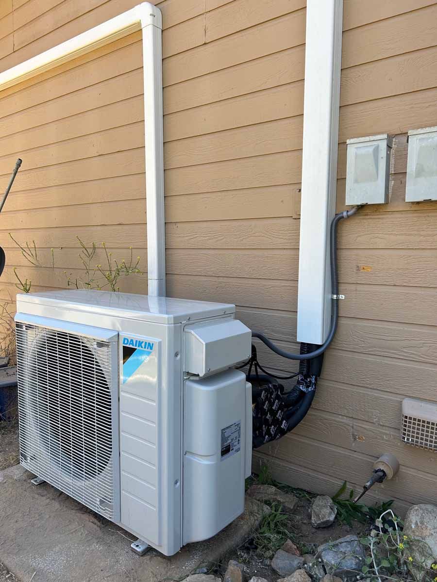 Outdoor HVAC condenser unit against a beige house wall with white piping and electrical boxes