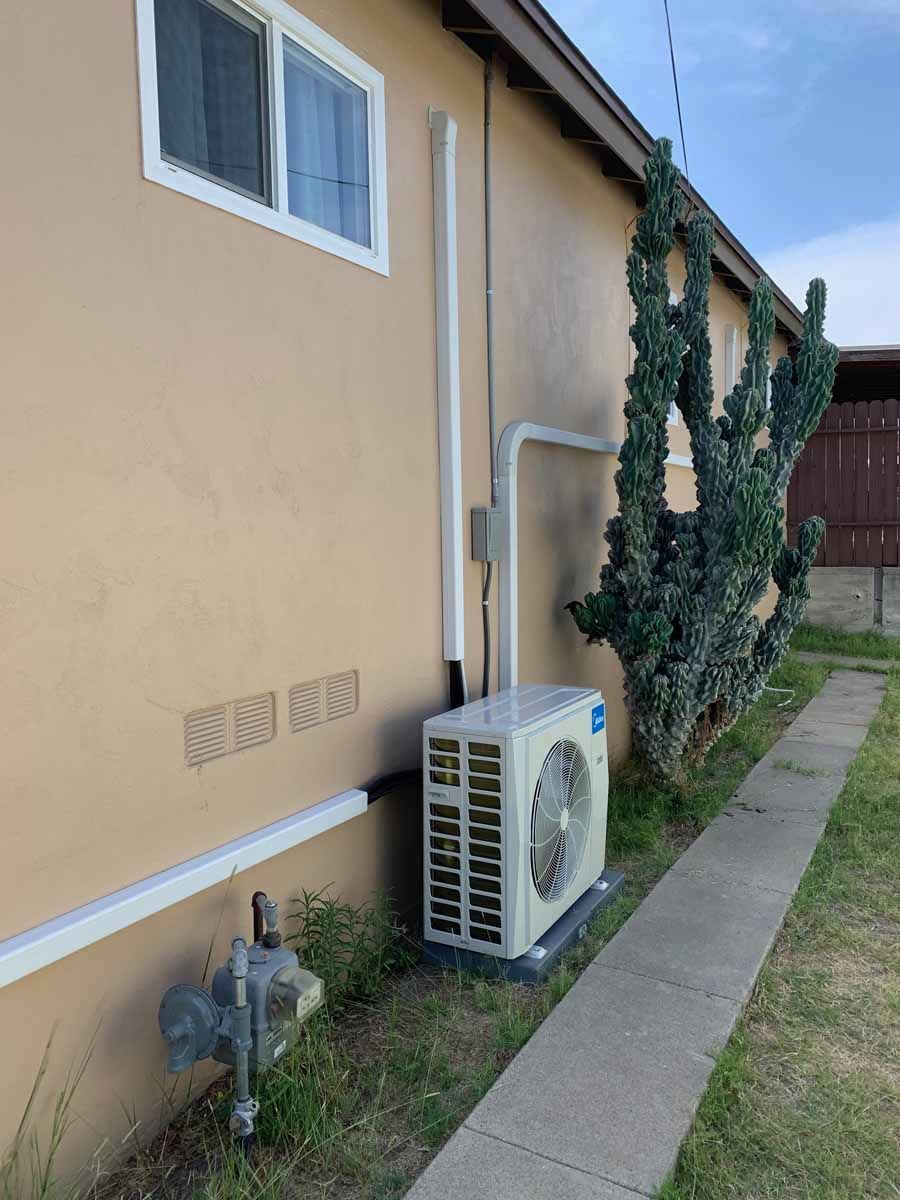 Side of a beige house with an air conditioner unit, utility pipes, and a narrow concrete walkway by ivy-covered wall.
