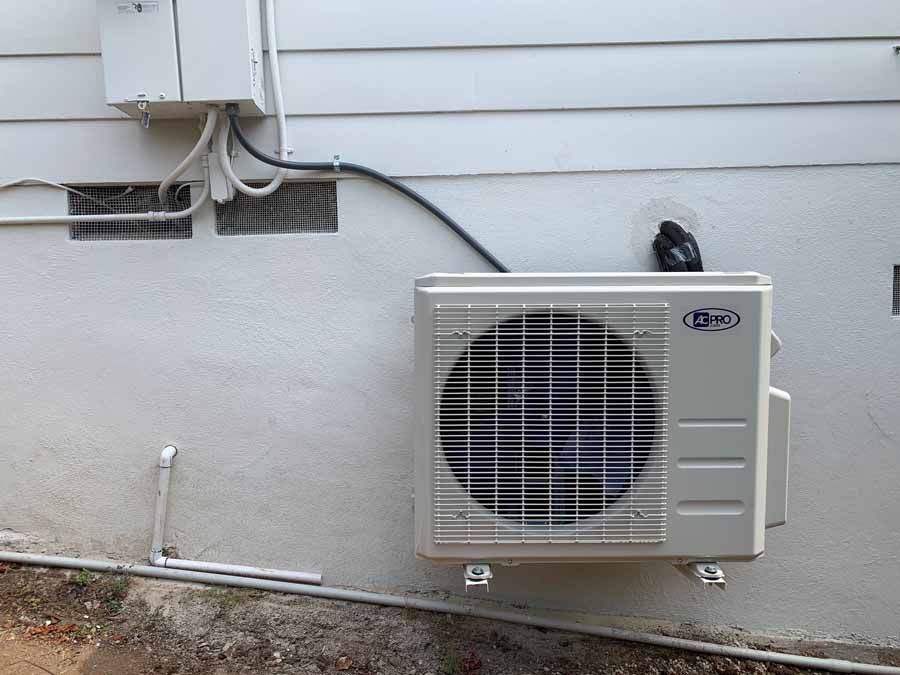 Outdoor air conditioner unit mounted on a house wall with visible cables and siding