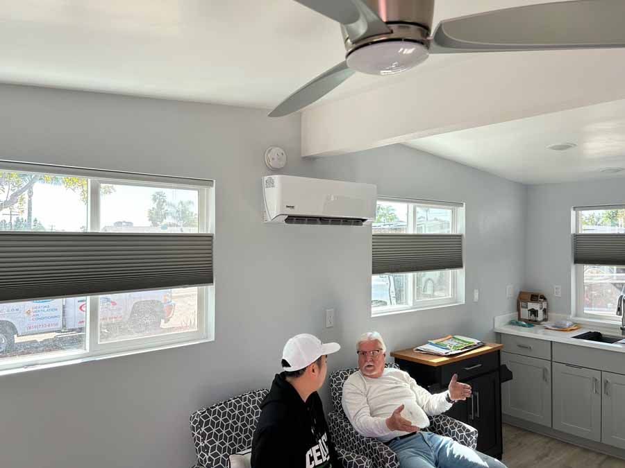 Bright living room with two people seated, ceiling fan, wall-mounted AC, and sunlight through windows