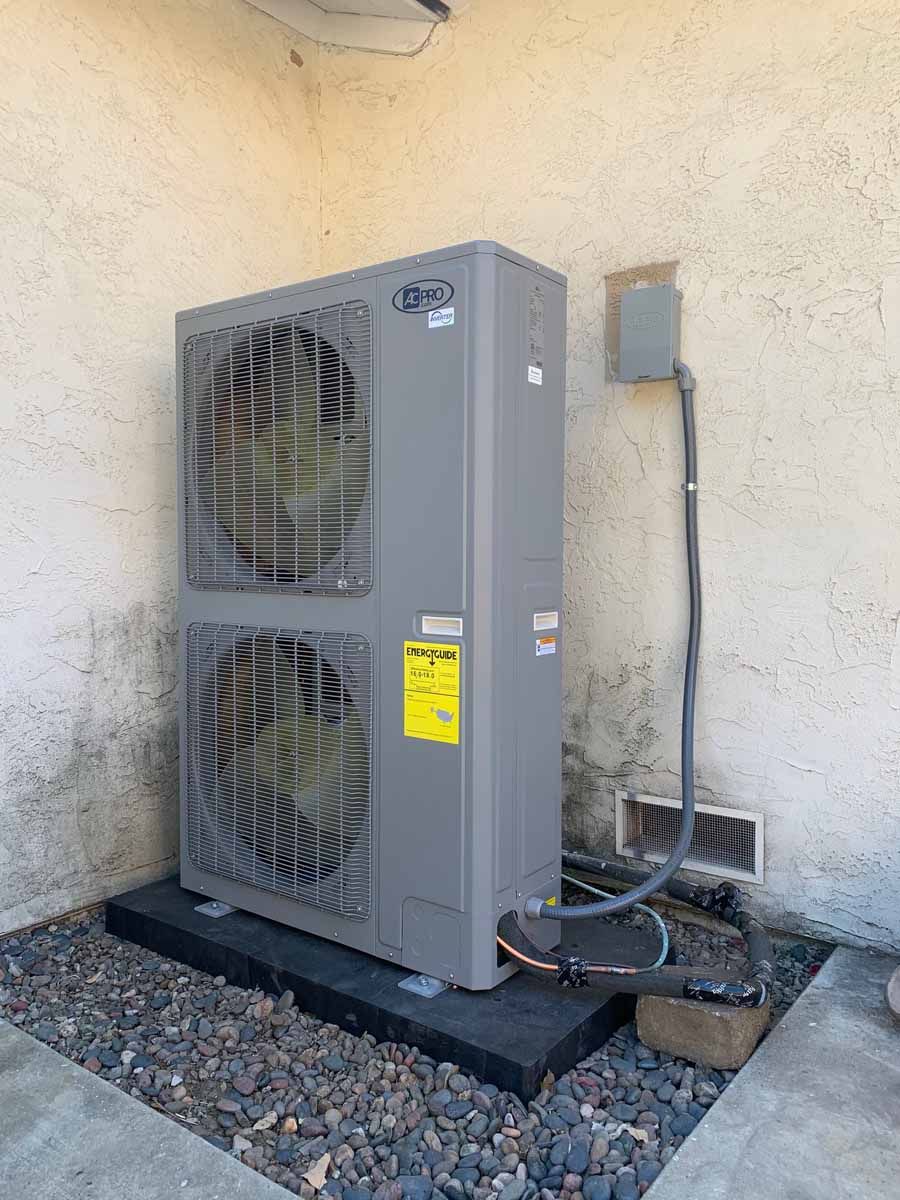 Outdoor air conditioner unit beside a beige wall, with piping and gravel ground.