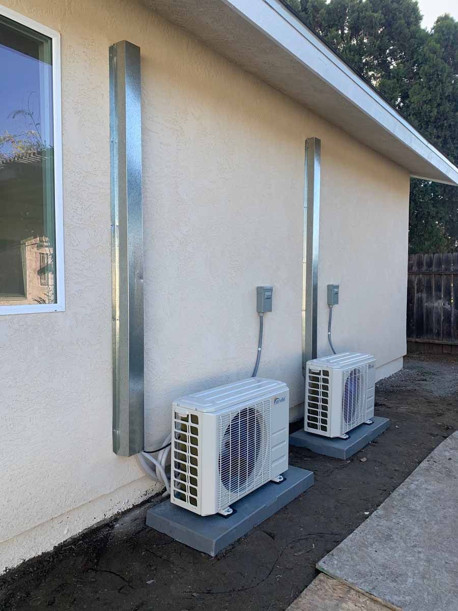 Two outdoor AC units beside a beige house wall on concrete pads, with vertical conduit pipes.
