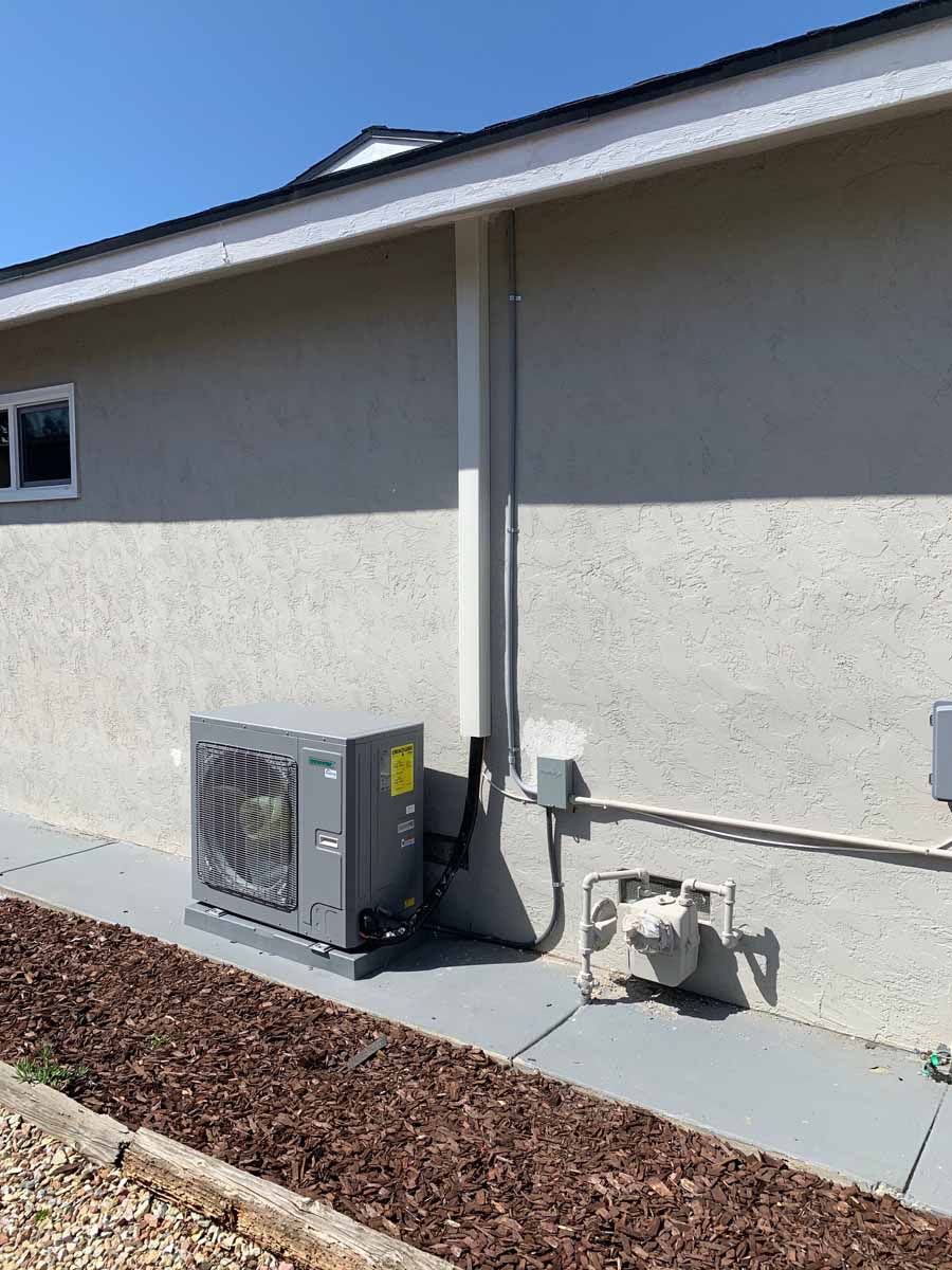 Outdoor HVAC unit and utility equipment beside a stucco house wall under a roof overhang.