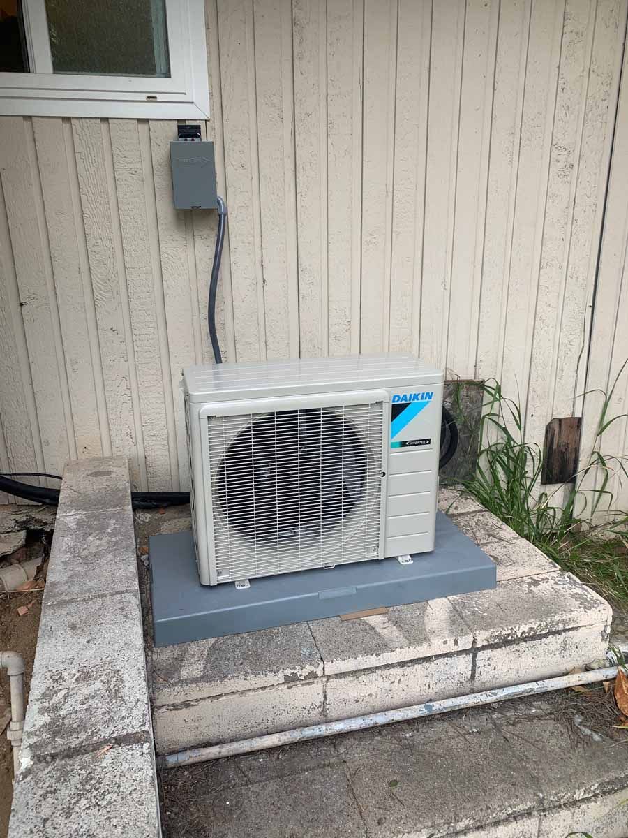 Outdoor air conditioner unit beside a beige wall on a concrete pad, with a window above.