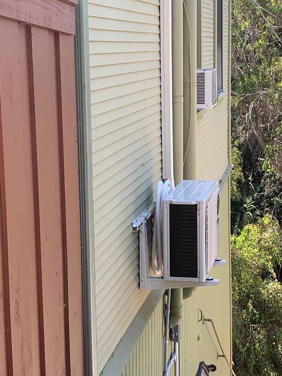 Wall-mounted air conditioner on the side of a beige house near brown siding and shrubs.