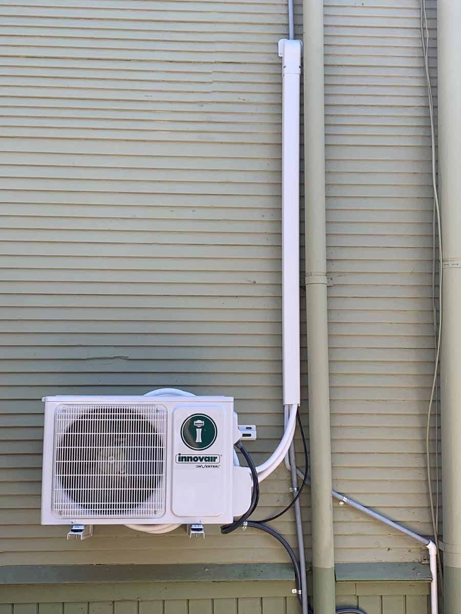 Outdoor wall-mounted air conditioner unit with white piping on a beige siding wall