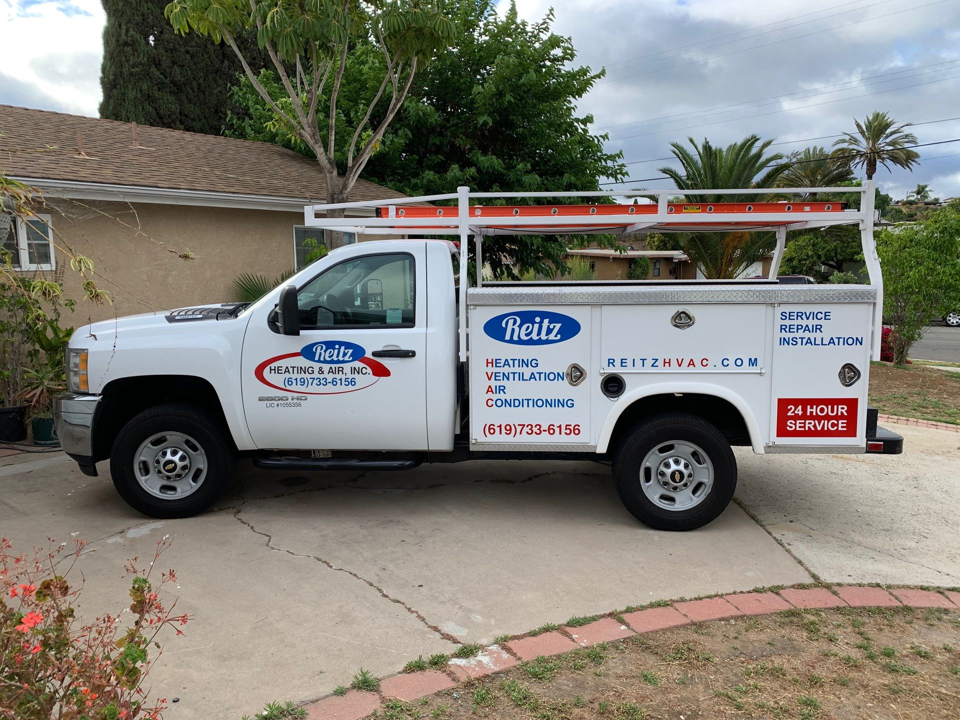 White service pickup truck with ladders parked in a driveway beside a house and palm trees.
