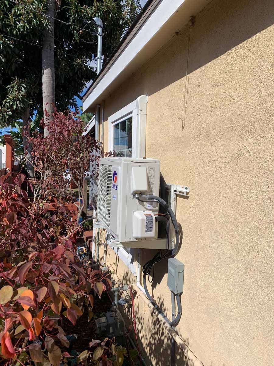 White outdoor utility meter box on a beige house wall beside red shrubs and trees.