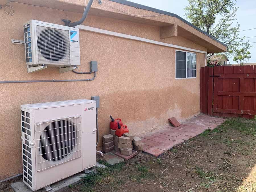 Two HVAC units mounted and on the ground beside a tan stucco house, with a red fence and yard