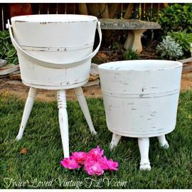 Two white vintage metal buckets on short legs, one tall and one short, with pink flowers on grass.