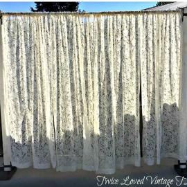 Lace curtain panel hanging outdoors in front of a window or wall, with sunlight and shadows visible