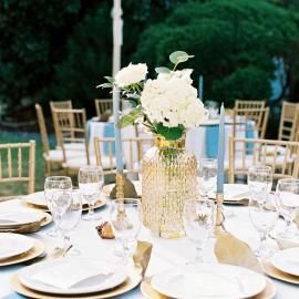 Elegant outdoor table with white dishes, gold chairs, and white floral centerpieces.
