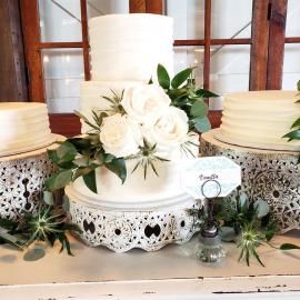 White tiered wedding cake with white roses and greenery on lace-trimmed stands in a rustic setting