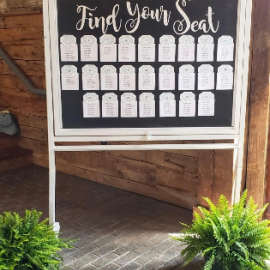 “Find Your Seat” wedding seating chart on a white-framed board with potted greenery at the base.