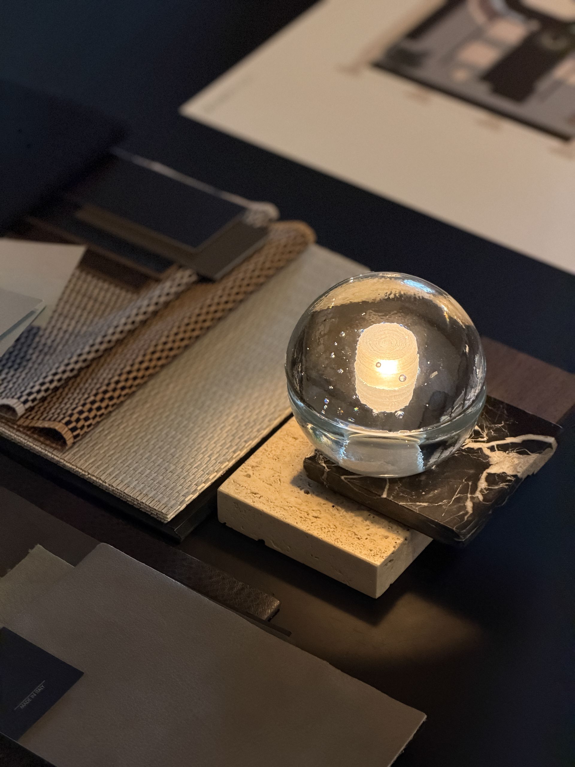 Glass orb on a textured display stand beside fabric samples and papers on a dark table.