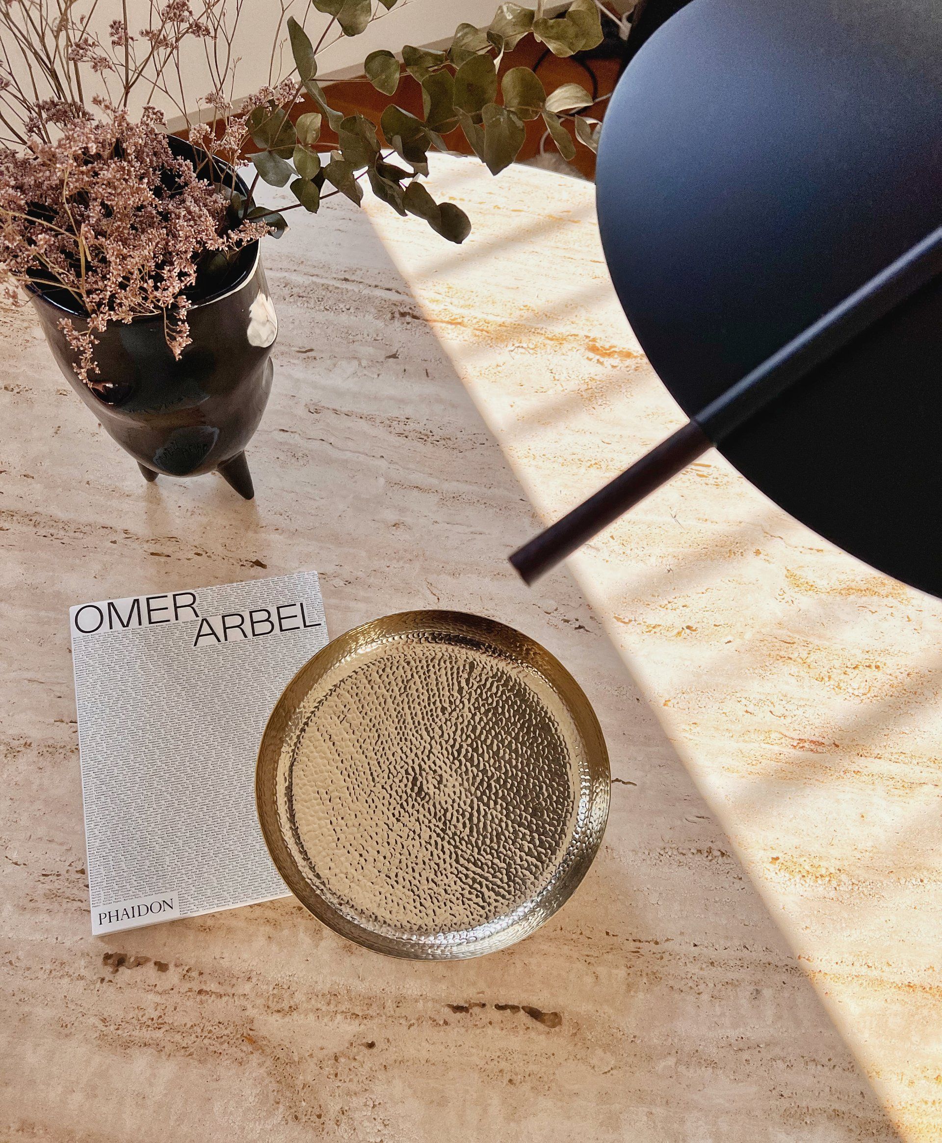 Gold tray, book, and dried flowers on a travertine, textured surface with a black table lamp in the corner.