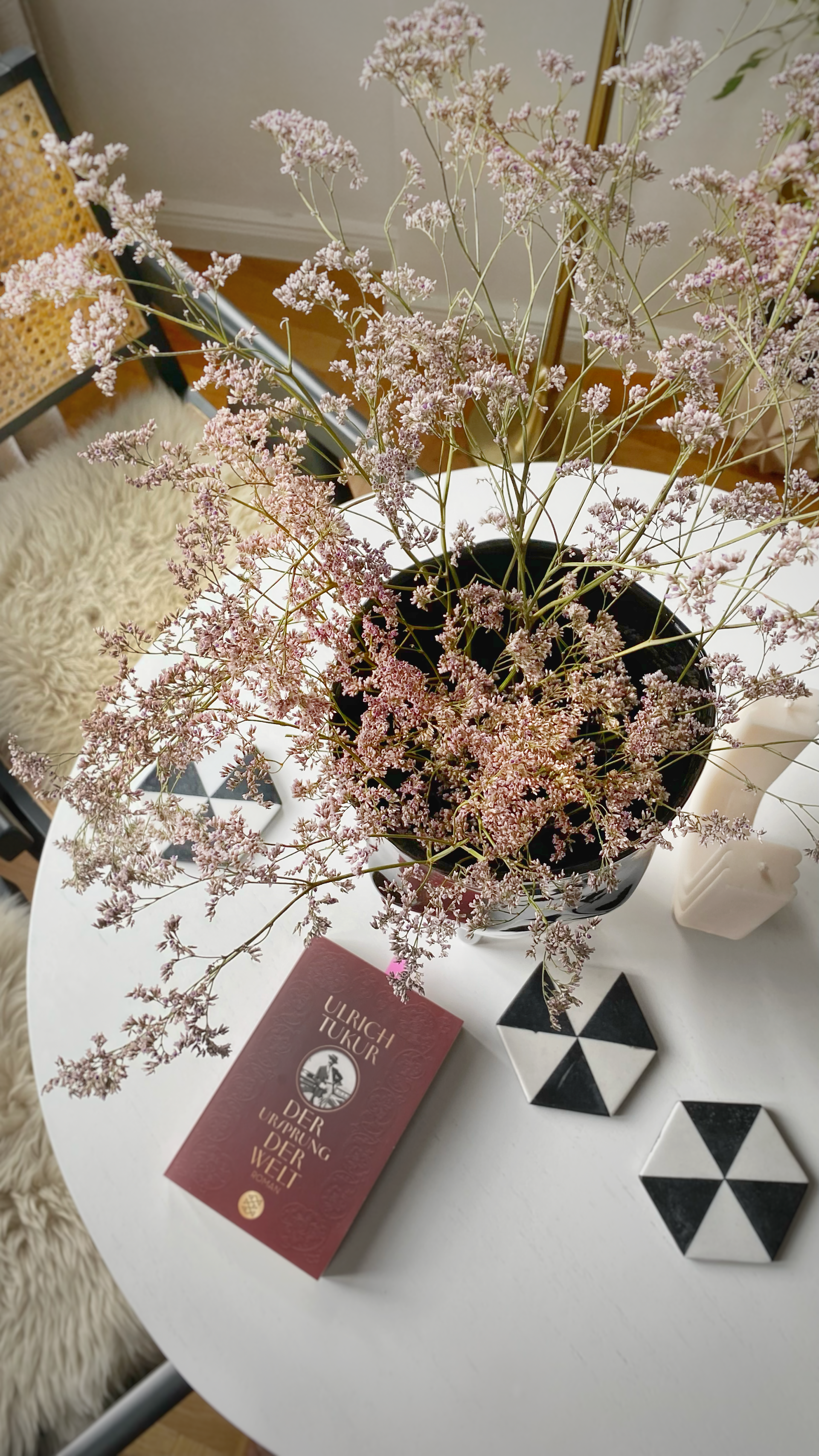 Pink flowers in a black vase on a white table, next to a book and geometric coasters.