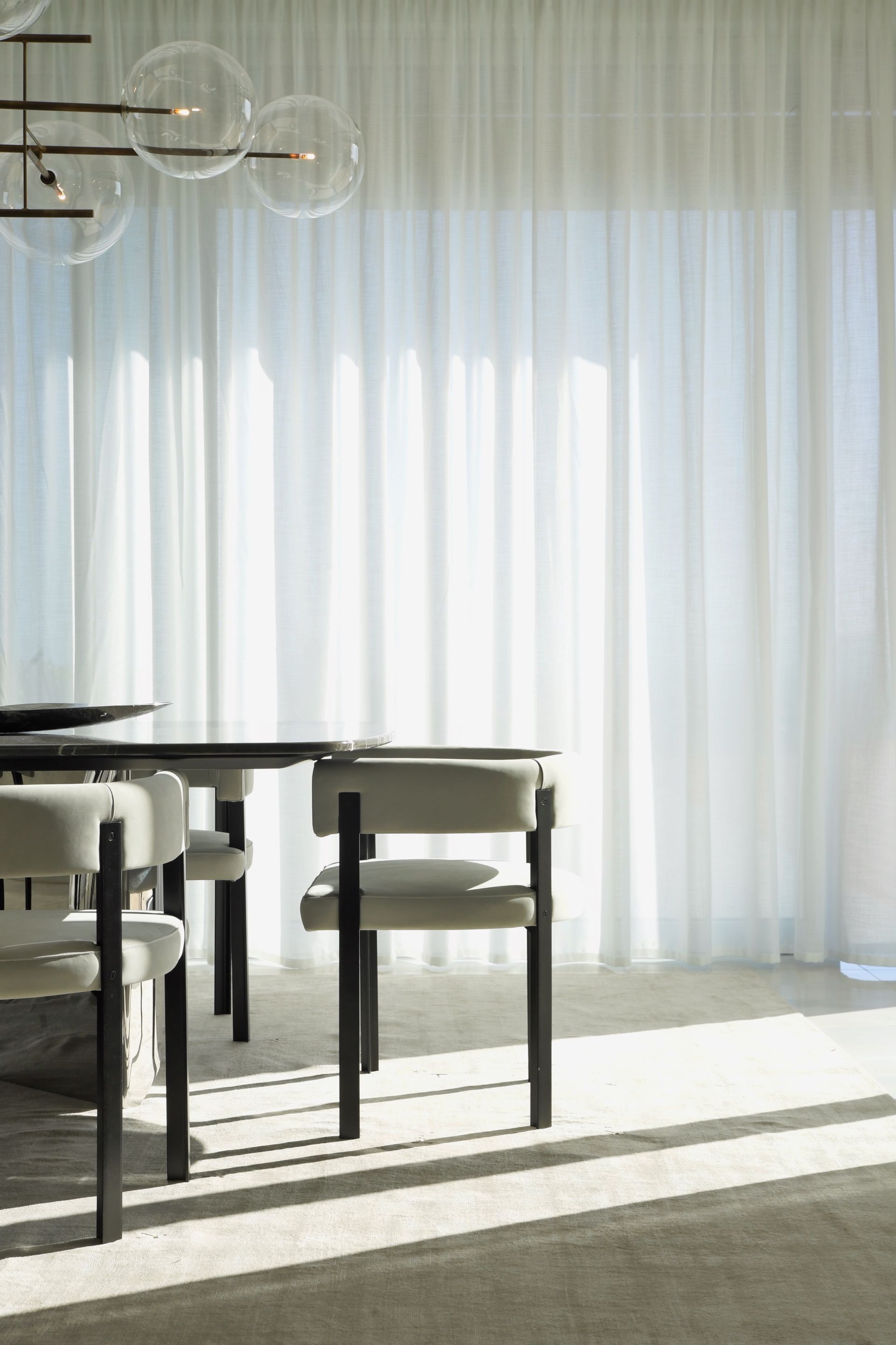 Dining room with table and chairs, sheer white curtains, and a modern chandelier. Sunlight streams in.