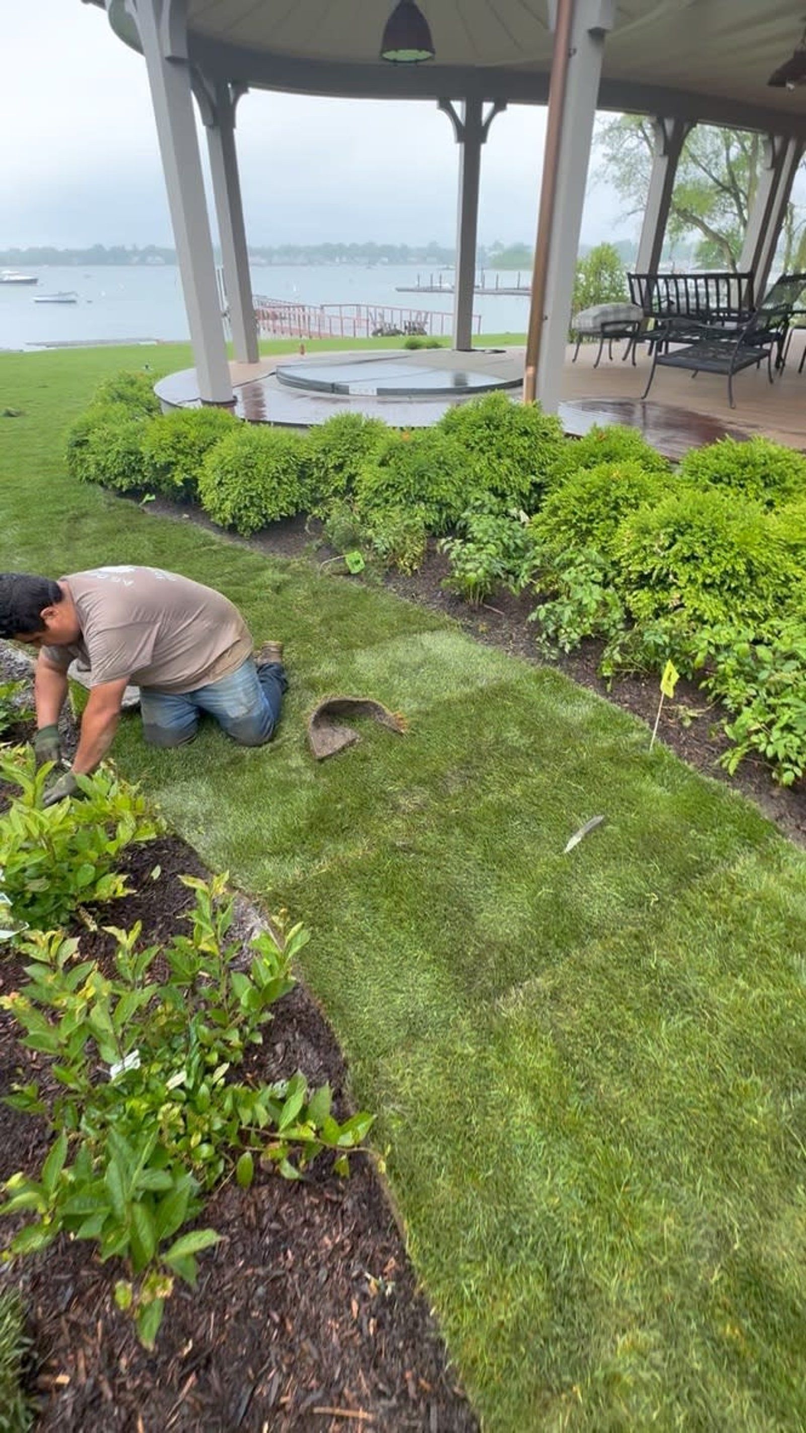 Gardener kneeling to trim shrubs beside a pavilion overlooking the water