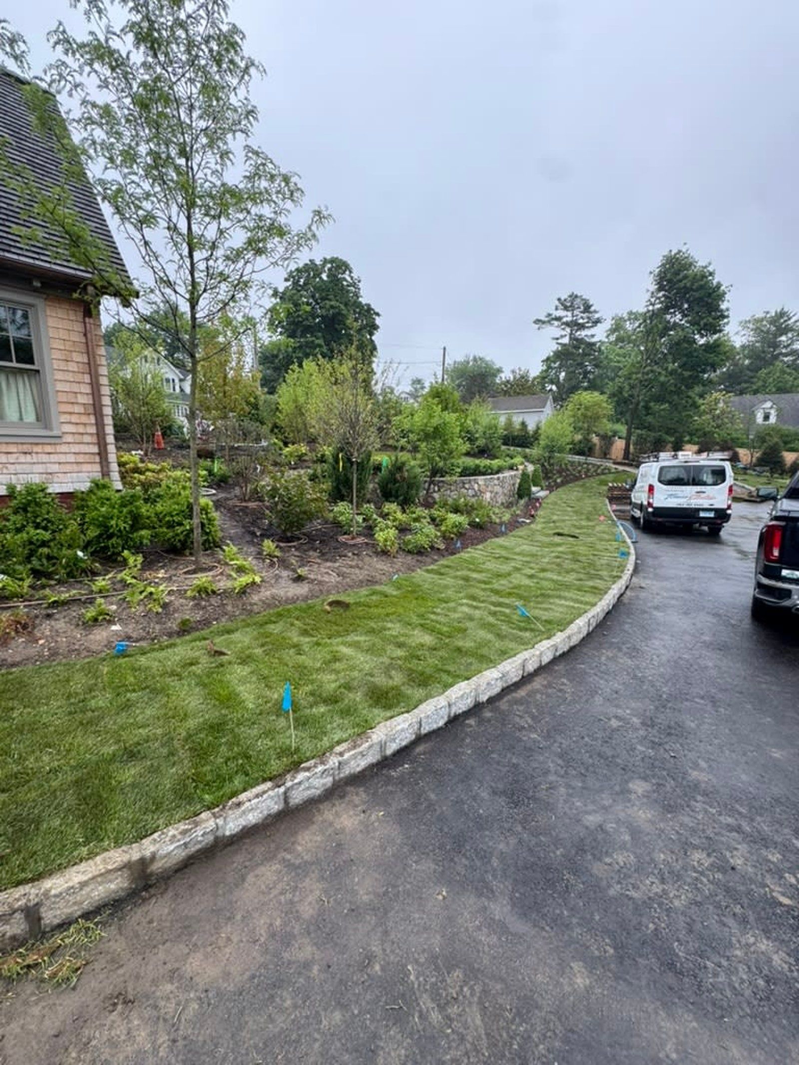 Curved driveway beside landscaped greenery and a brick building, with parked cars on a wet road under overcast skies