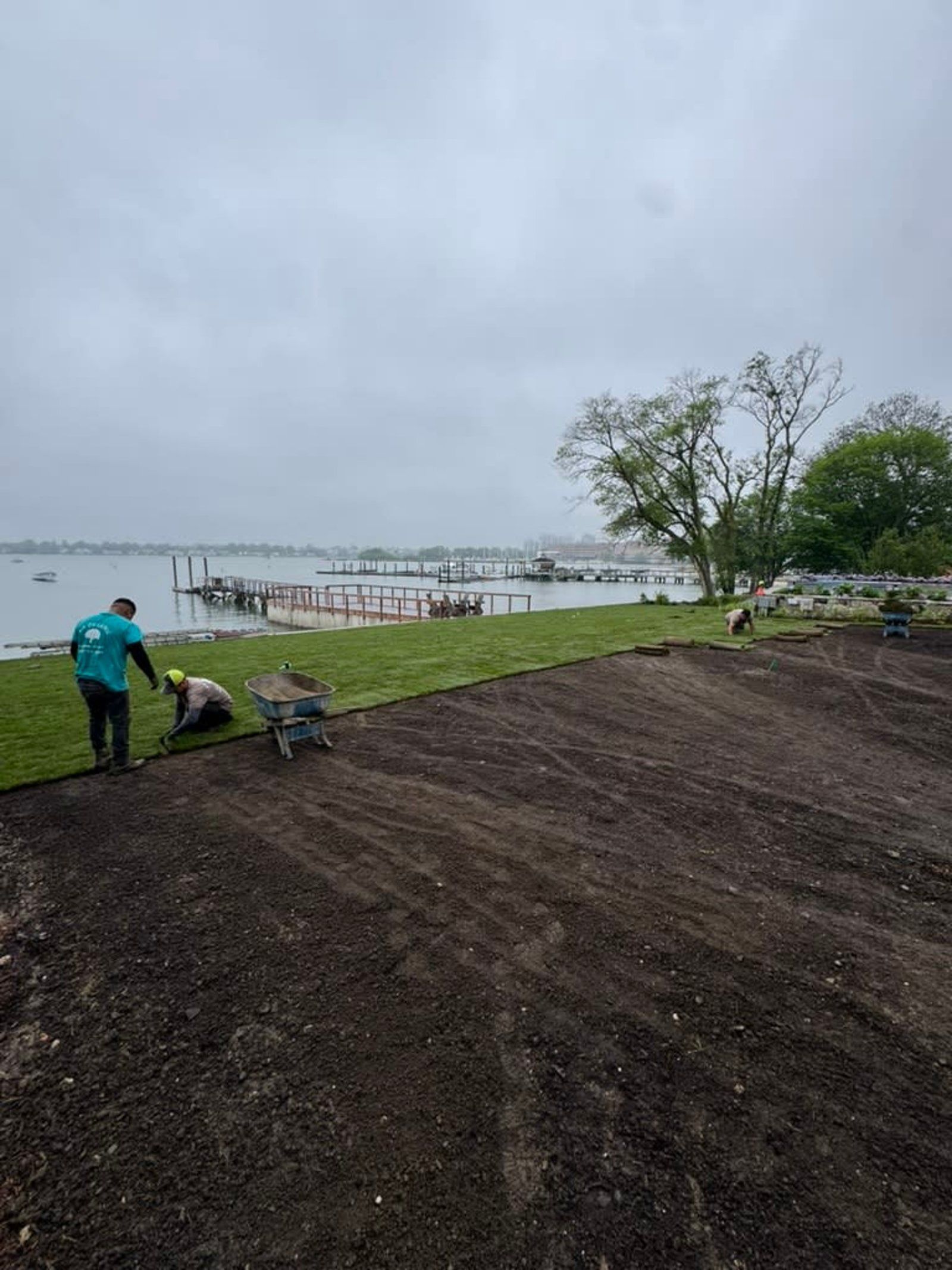 Two people work beside a muddy shoreline with a dock and calm water under a cloudy sky.
