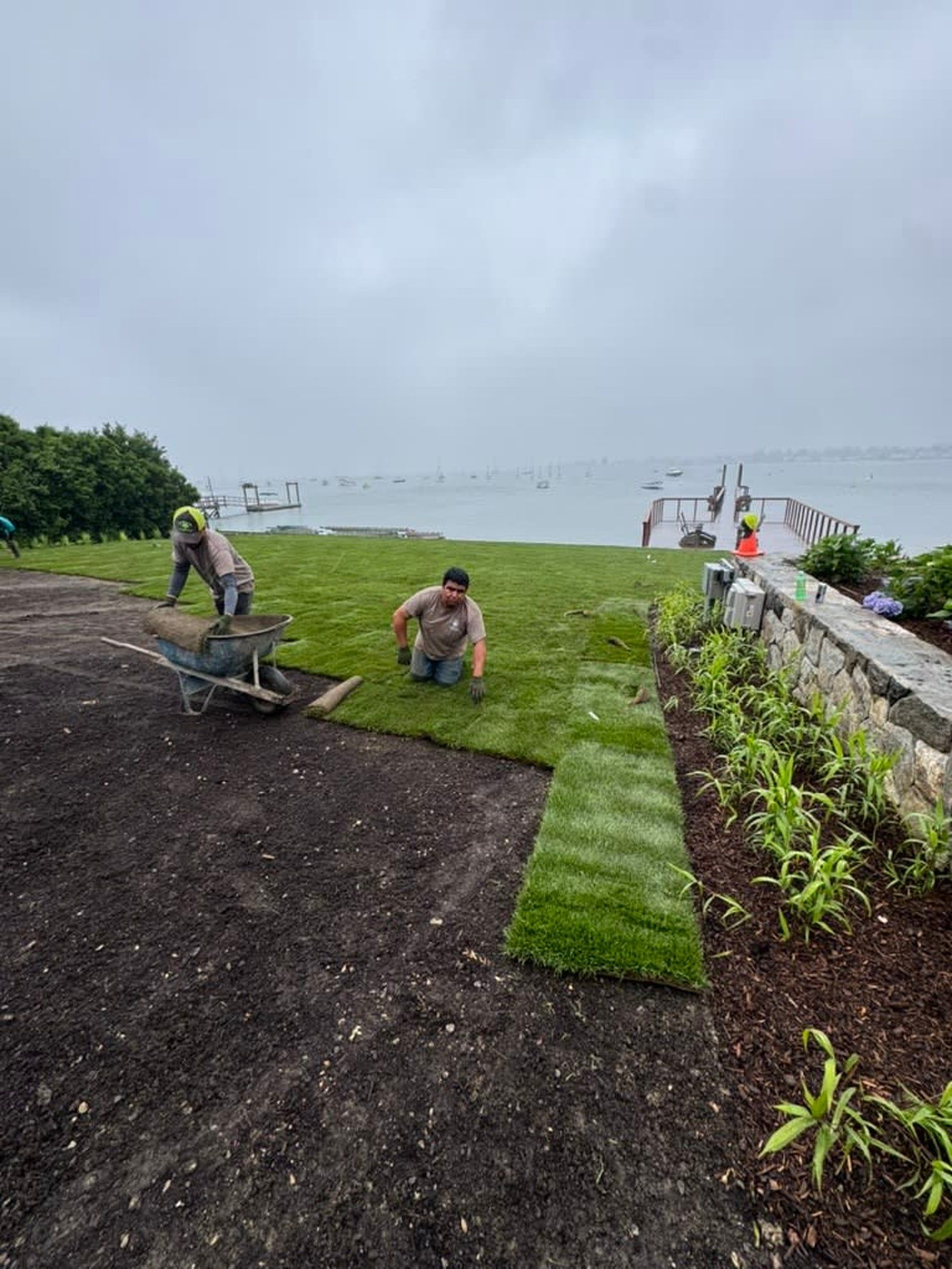 Workers laying sod on a grassy coastal slope under a gray sky, with a path and ocean view nearby