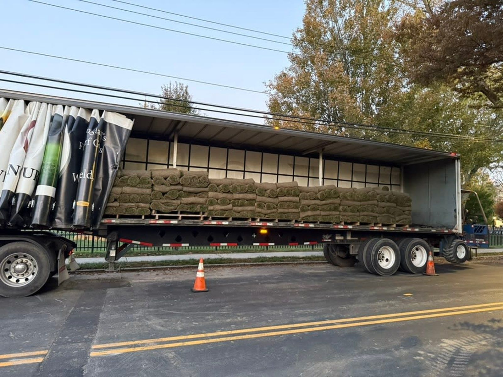 Flatbed semi-trailer carrying stacked hay bales parked on a roadside, with an orange cone nearby.