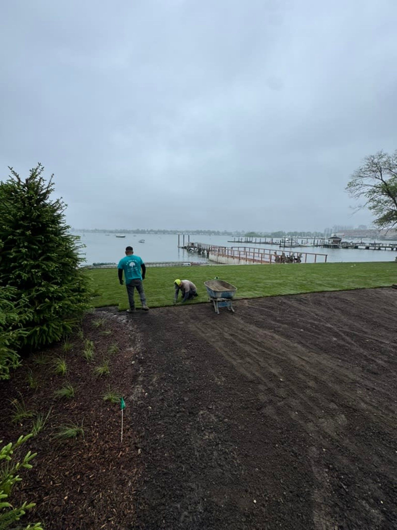 Two people gardening by a waterfront lawn under a cloudy sky, with boats in the distance.