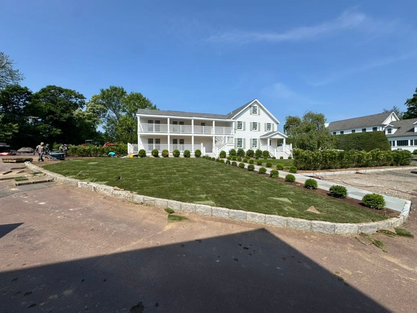 White two-story house with wraparound porch on a landscaped hill under a blue sky