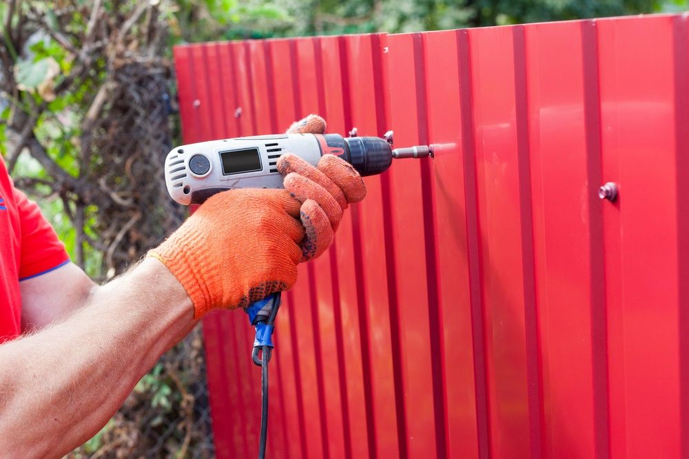 A Man Is Using A Drill To Drill A Hole In A Red Fence — Ballina Best Fencing In Northern Rivers, NSW