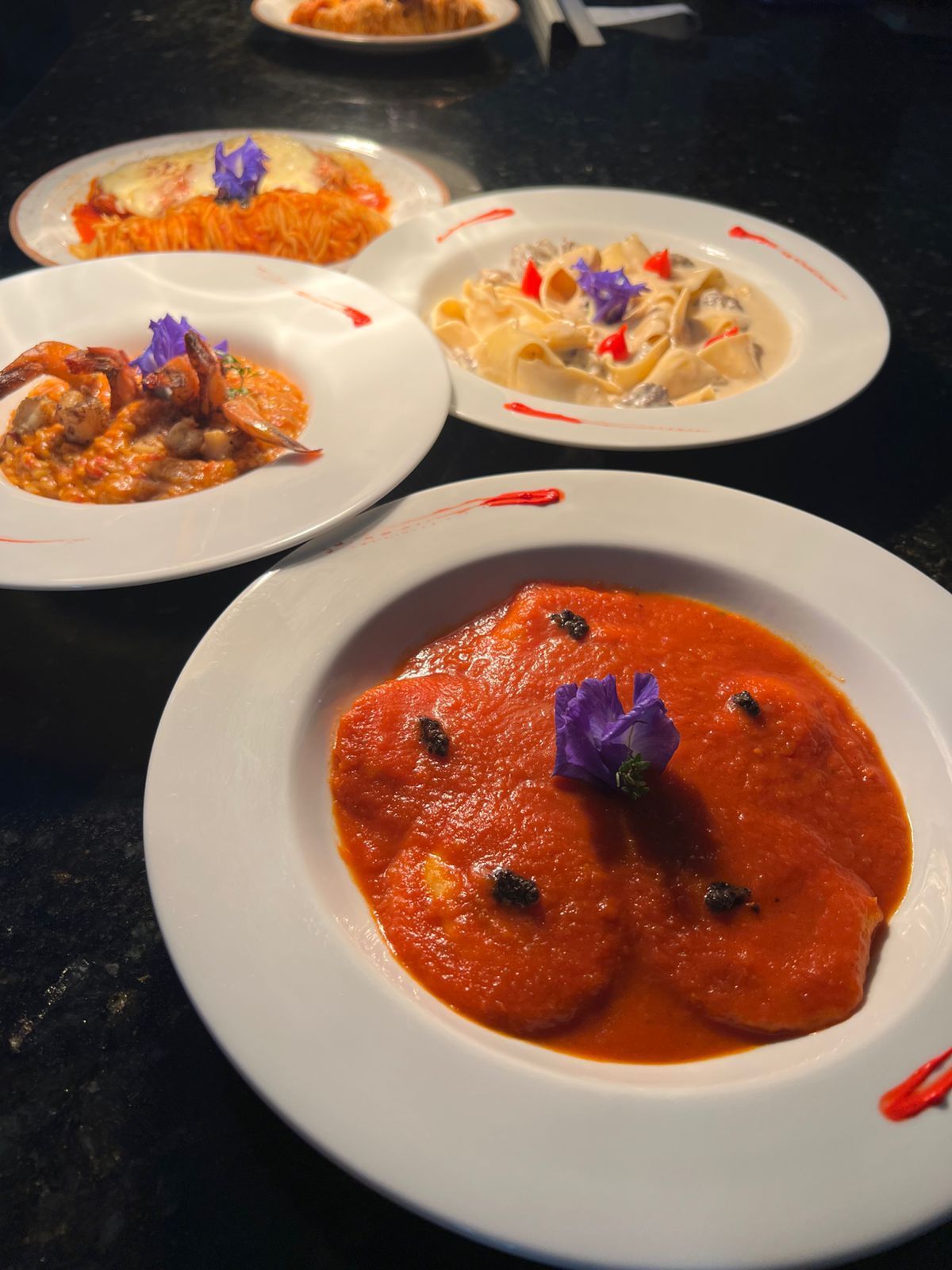 Quatro pratos de massa: opções à base de tomate, cremosas e frutos do mar, decorados com flores em uma mesa escura.Pousada Villa Italiana