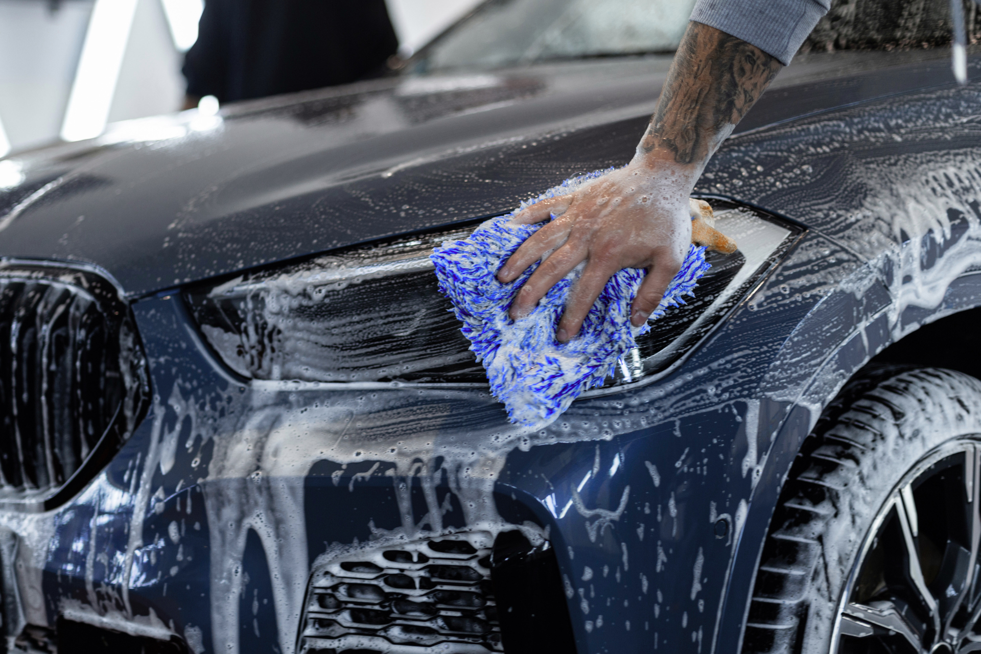 Hand washing a black car’s front headlight and hood with a blue sponge and soapy water