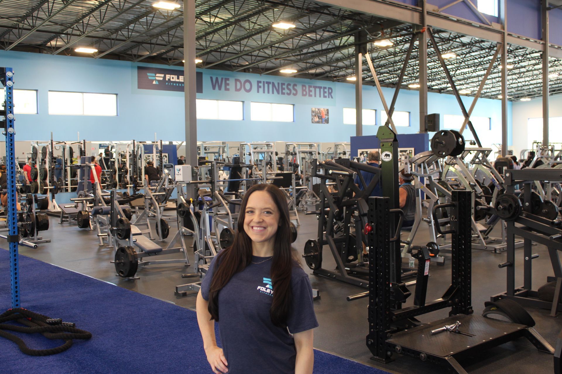 Woman in teal shirt smiles at gym; exercise equipment and people in background.