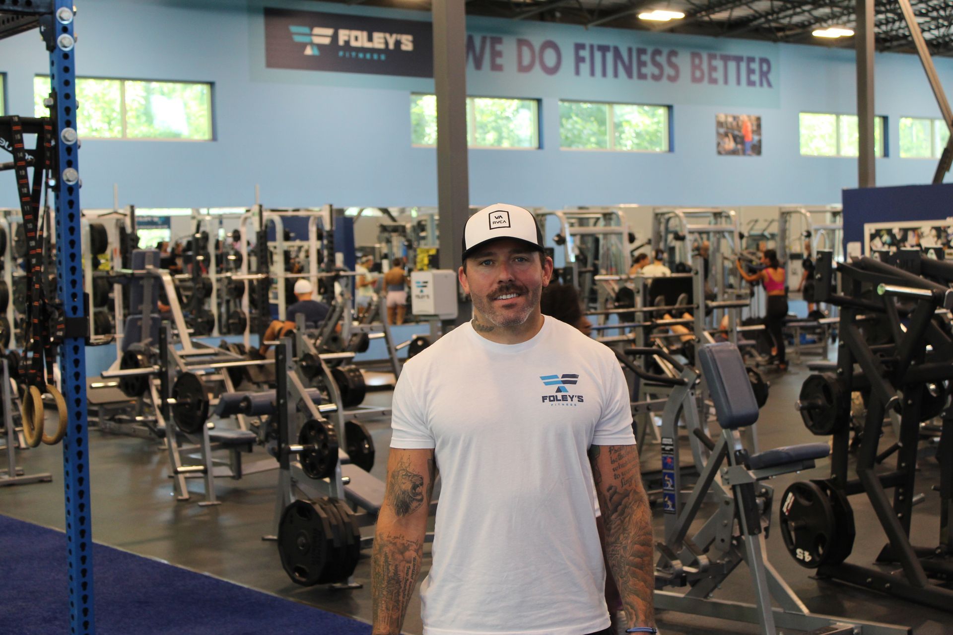 Woman in teal shirt smiles at gym; exercise equipment and people in background.