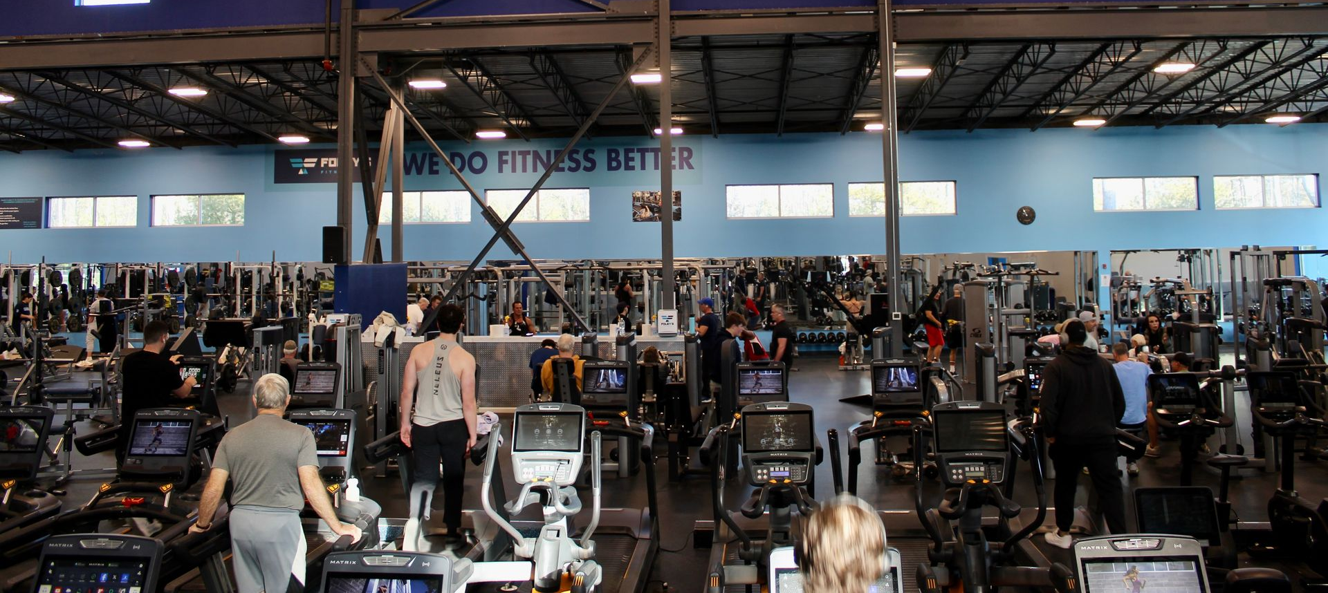 People exercising on machines in a gym with high ceilings, windows, and exercise equipment.