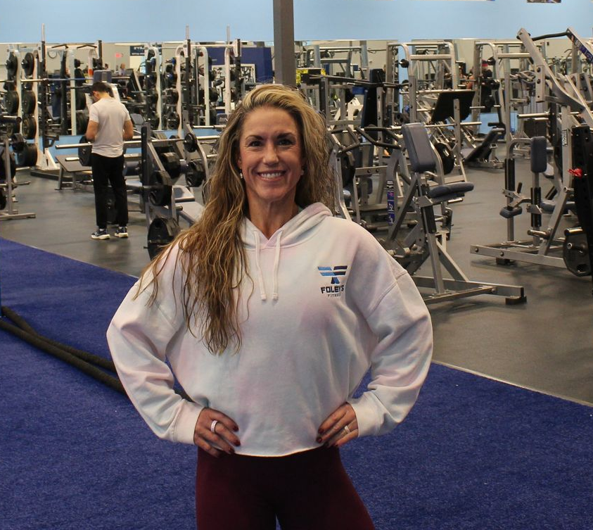 Woman in white sweatshirt at a gym, hands on hips, smiling. Many weight machines in the background.