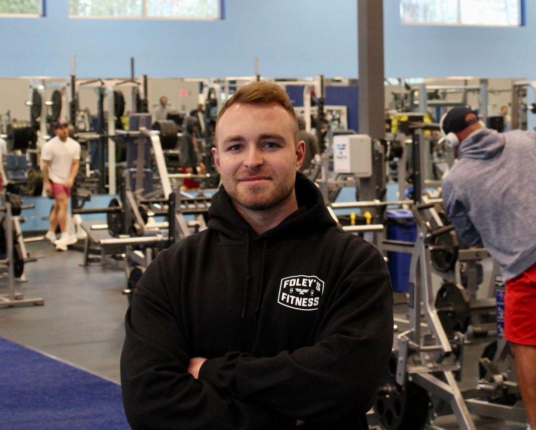 Man in black hoodie with arms crossed, in a gym. Other people and equipment in background.