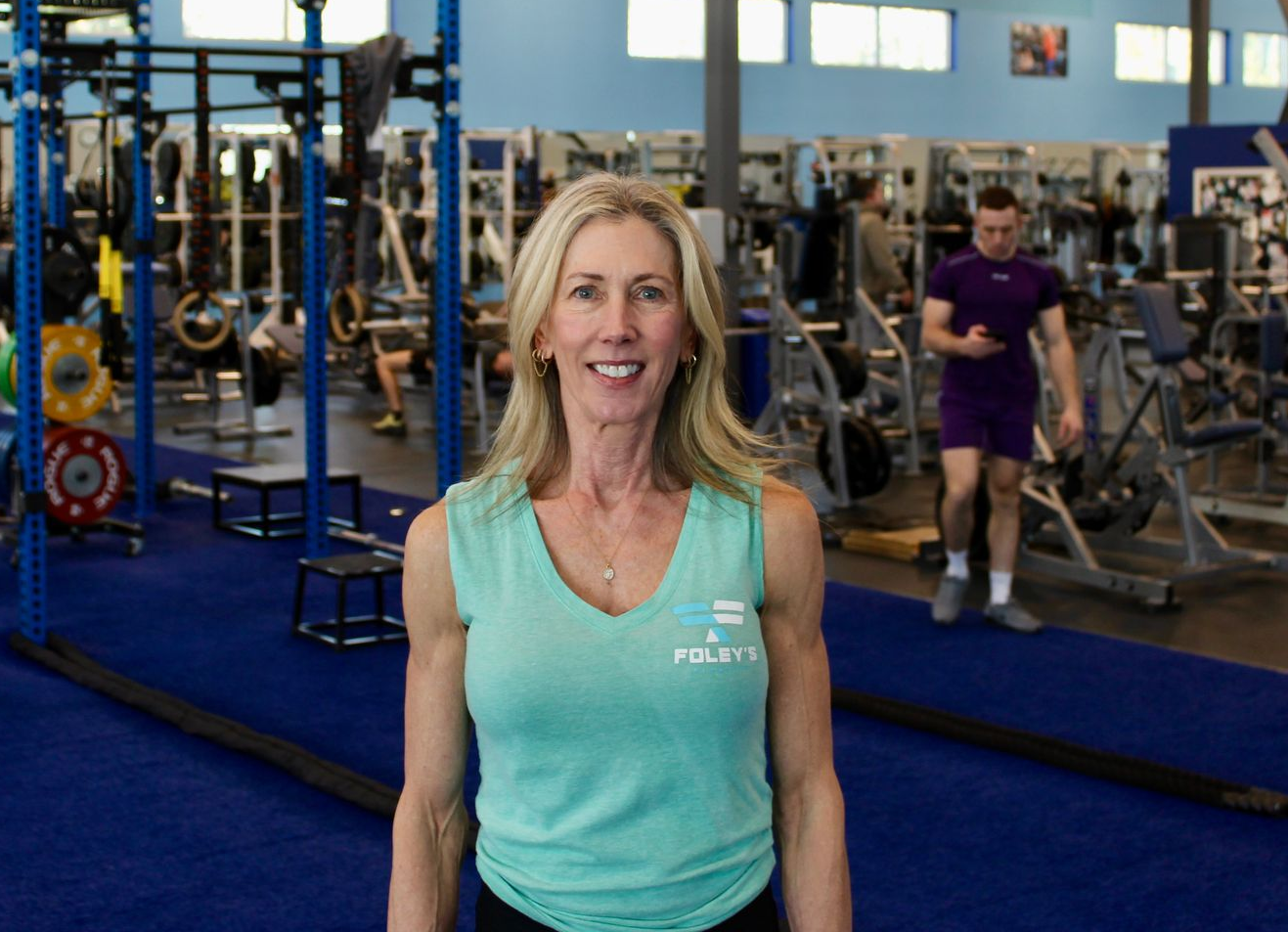 Woman in teal shirt smiles at gym; exercise equipment and people in background.
